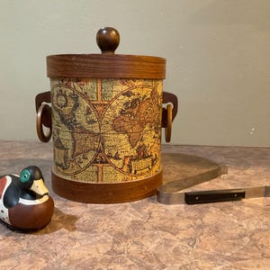 May include: A wooden ice bucket with a world map design. The bucket has two handles and a lid. A small duck figurine is in the foreground.