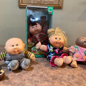 May include: Four Cabbage Patch Kids dolls, one in a red and green holiday outfit, one in a black and pink floral dress, one in a blue and green striped outfit, and one in a pink and white floral dress. The dolls are all sitting on a brown surface.