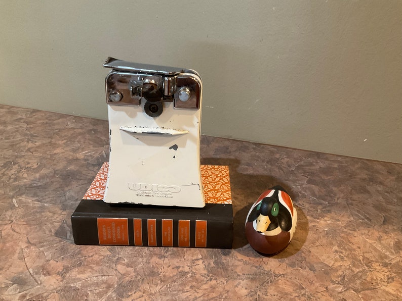 Peut inclure: Un ouvre-bo&icirc;te blanc avec une poign&eacute;e argent&eacute;e et un livre noir et orange avec le texte "BRIGHTON" sur le dos. Une petite figurine de canard brun et vert se trouve &agrave; droite de l'image.