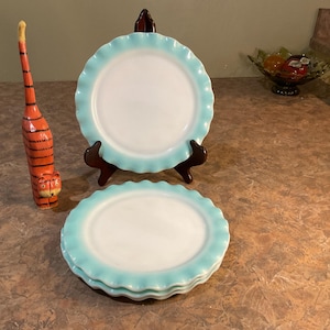 May include: A stack of white plates with a light blue scalloped edge. One plate is displayed on a brown stand. A decorative orange and black tiger figurine stands to the left of the plates. The plates are on a brown countertop.
