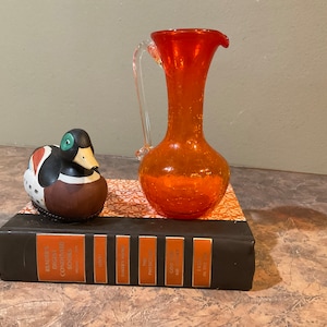 May include: A small orange glass pitcher with a clear handle sits on top of a brown book with a white and orange patterned cover. A small ceramic duck figurine is on the book to the left of the pitcher.
