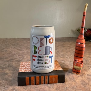 May include: A ceramic beer stein featuring the text Oktoberfest München 21. September bis 6. Oktober 1991, decorated with colorful icons. It sits on a black and orange book next to a tall, slender, orange and black hand-carved wooden cat figurine.