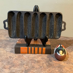 Peut inclure: Un moule à pain de maïs en fonte avec six moules individuels. Le moule est noir et a une surface texturée qui ressemble à des grains de maïs. Le moule est posé sur un livre avec une couverture brune.