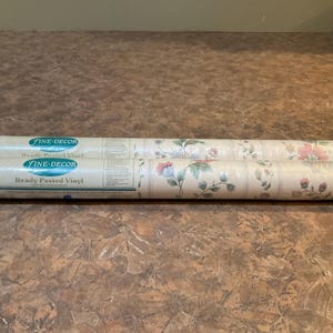 May include: Two rolls of Fine-Decor Ready Pasted Vinyl wallpaper. The wallpaper features a floral pattern with pink and green flowers on a cream background. The rolls are lying on a brown surface.