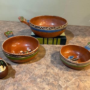 May include: Three terracotta bowls with painted designs. The bowls have handles and are decorated with blue, green, and white accents. The bowls are sitting on a brown surface.