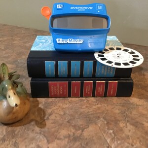 Puede incluir: Un juguete View-Master azul con un carrete 3D Overdrive Plus, un carrete View-Master blanco y dos libros de tapa dura negros apilados uno encima del otro. Un pequeño macetero de cerámica con forma de pato con una planta suculenta en primer plano.