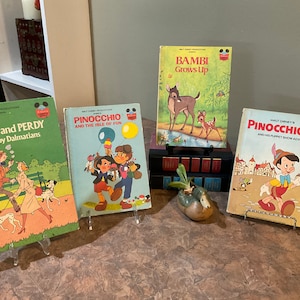 May include: Four vintage Disney children's books on display stands. The books are titled "Pongo and Perdy Two Happy Dalmatians", "Pinocchio and the Isle of Fun", "Bambi Grows Up", and "Walt Disney's Pinocchio and His Puppet Show Adventure".
