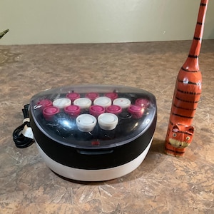 Op de afbeelding: Een zwart-witte haarroller set met roze en witte rollers. Een gesneden houten tijgerbeeldje met oranje en zwarte strepen staat in de buurt. Een decoratieve kom met rood fruit staat op de achtergrond.