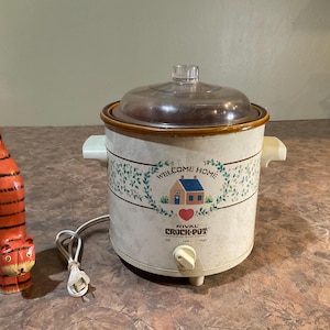 May include: A Rival Crock-Pot slow cooker with a cream-coloured exterior, a floral border, a house graphic, and the text "WELCOME HOME" above a red heart. Beside it stands a tall, slender, orange and black striped wooden cat figurine.