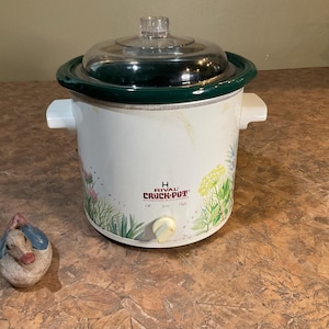 Op de afbeelding: Een witte Rival Crock-Pot slowcooker met een doorzichtig glazen deksel en een donkergroene rand. De slowcooker heeft bloemmotieven en een bedieningsknop. Links van de slowcooker staat een klein decoratief beeldje.