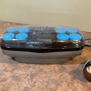 Op de afbeelding: Een Conair-set met warme rollers met een doorzichtig plastic deksel. De rollers zijn zwart met blauwe plastic doppen. De set staat op een bruine en beige betegelde ondergrond. Een klein bruin en groen eendje staat op de voorgrond.