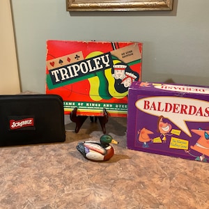 May include: A collection of board games including Tripoley, Balderdash, and Scrabble. The Tripoley game box is red, green, and yellow with the text "Tripoley" in white. The Balderdash game box is purple with the text "Balderdash" in white. The Scrabble game is in a black carrying case with the text "Scrabble" in red. A wooden duck figurine is also in the image.