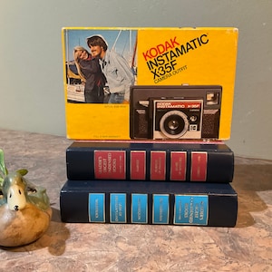 以下が含まれることがあります： 黄色い箱に入ったビンテージのKodak Instamatic X35Fカメラ。箱にはボートに乗ったカップルが写っています。カメラは黒で、レンズは銀色です。箱には「Kodak Instamatic X35F Camera Outfit」と書いてあります。カメラは、背表紙に「Reader's Digest Condensed Books」と書かれた2冊の青いハードカバーの本の上に置かれています。