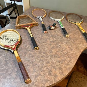 May include: A collection of vintage tennis rackets with wooden frames and various grip styles. The rackets are arranged on a table, showcasing their aged appearance and design. A small decorative duck figurine is also present.