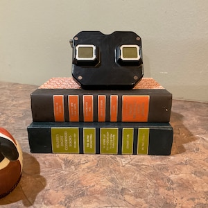 Op de afbeelding: Een zwarte vintage 3D-viewer ligt op twee boeken. Het bovenste boek is bruin met gouden letters die "Reader's Digest Condensed Books" lezen. Het onderste boek is zwart met gouden letters die "On the Trail of the Ghost Fox" lezen.