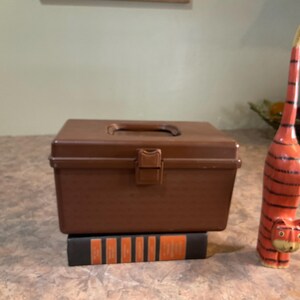 May include: A brown plastic toolbox with a handle and clasp sits on a decorative block with orange stripes. A carved wooden tiger figurine with orange and black stripes stands nearby.