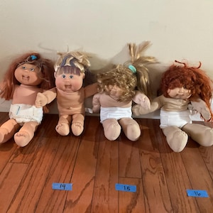 Op de afbeelding: Vier vintage Cabbage Patch Kids poppen met verschillende haarkleuren, waaronder rood, blond en bruin. De poppen dragen luiers en hebben geschilderde gezichten. Ze zijn gerangschikt op een houten vloer.