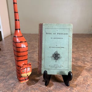May include: A tall, hand-carved wooden cat figurine painted orange with black stripes and a light-colored tail tip, standing next to an antique book titled A Book of Problems in Arithmetic by Geo A. Walton and Francis Cogswell, Boston 1877.