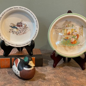 Puede incluir: Dos platos de guardería vintage de Beatrix Potter, uno con Peter Rabbit y el otro con un lechón con un plato de carne asada. Los platos están en pequeños soportes de madera. Una pequeña figura de pato está en primer plano.