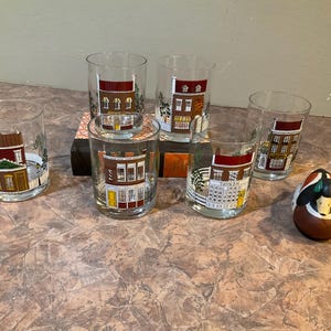 May include: Six clear glass tumblers with painted scenes of houses and a small wooden duck figurine. The tumblers are decorated with red, brown, and white paint.