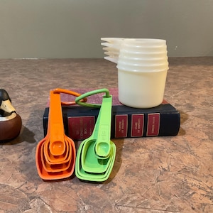 Peut inclure: Un ensemble de quatre cuillères doseuses, deux orange et deux vertes, avec une pile de cuillères doseuses blanches en arrière-plan. Les cuillères doseuses sont posées sur un livre.