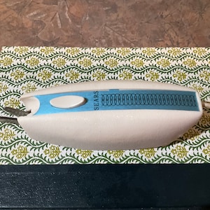 Könnte beinhalten: Weiße elektrische Stoffschere mit blauem Griff und schwarzem Kabel. Die Schere hat eine Messhilfe oben und das Wort "SEARS" ist seitlich aufgedruckt.