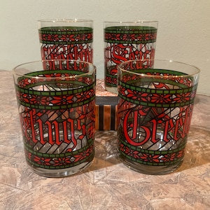 May include: A set of four clear glass tumblers featuring a stained-glass style pattern in red and green. The design includes a decorative border of poinsettia-like flowers and the red text Season's Greetings wrapped around the exterior of each glass.