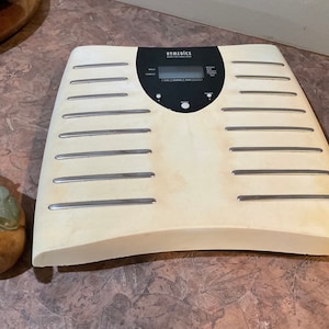 Peut inclure: Une balance numérique blanche avec un écran noir et le mot "HOMERICS" imprimé en haut. La balance a une bordure argentée autour des bords et un bouton argenté à l'avant.