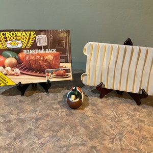 May include: A white ceramic roasting rack with a brown wooden stand. The rack is designed for use in both microwave and conventional ovens. The box for the rack is also pictured, featuring a photo of a roast on the rack. The text on the box reads "Microwave Roasting Rack".