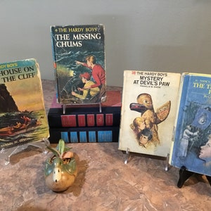Peut inclure: Quatre livres brochés vintage sur des supports d'exposition. Les livres sont tous des romans policiers, avec des titres comme "The House on the Cliff", "The Missing Chums", "Mystery at Devil's Paw", et "The Thirteenth Pearl".