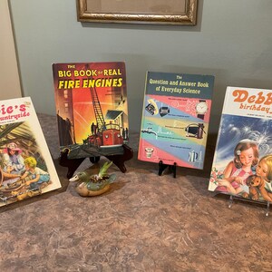 Puede incluir: Cuatro libros infantiles vintage sobre una mesa de madera. Los libros se titulan "Debbie's visit to the countryside", "The Big Book of Real Fire Engines", "The Question and Answer Book of Everyday Science", y "Debbie's birthday party".