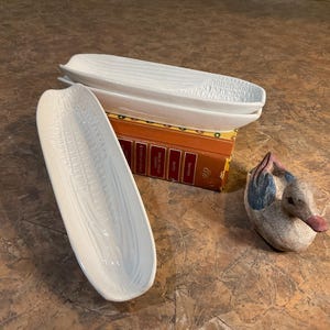 May include: Three white ceramic corn-shaped serving dishes are stacked on a book, with a decorative duck figurine nearby. The dishes have a textured surface, and the duck has blue and pink accents.