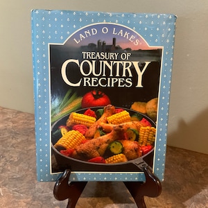 May include: A blue cookbook with a white pattern and the title "Treasury of Country Recipes" by Land O'Lakes. The cover features a photo of a skillet with chicken, corn, zucchini, and tomatoes.