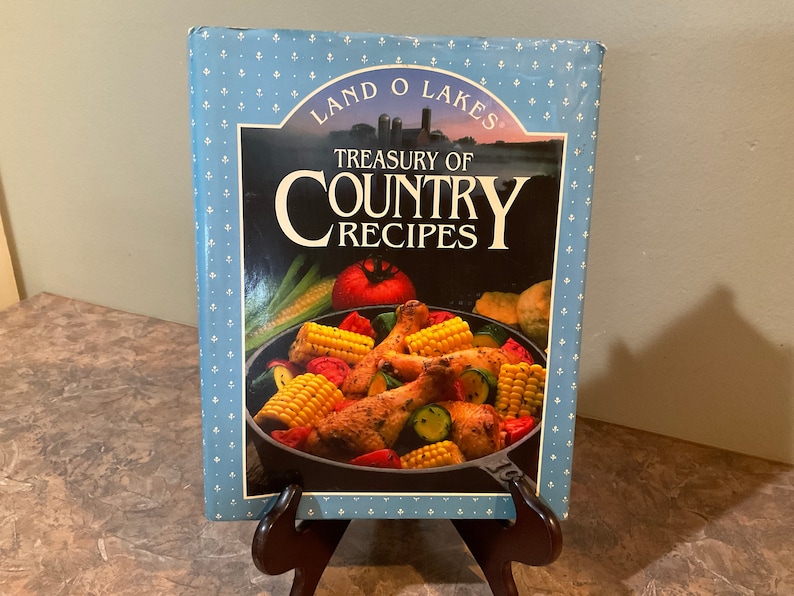 May include: A blue cookbook with a white pattern and the title "Treasury of Country Recipes" by Land O'Lakes. The cover features a photo of a skillet with chicken, corn, zucchini, and tomatoes.