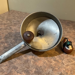 Puede incluir: Una cacerola de aluminio con un mango largo y un pomo de madera en la tapa. Una figura decorativa de pato de madera con detalles verdes, rojos y marrones se encuentra junto a la cacerola. La cacerola tiene un aspecto vintage y desgastado.