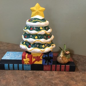 Puede incluir: Un tarro de galletas con forma de árbol de Navidad de cerámica con una estrella amarilla, decorado con bastones de caramelo y adornos. El árbol se asienta sobre una base de regalos envueltos y libros. A la derecha hay una pequeña maceta de pato.