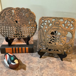 Puede incluir: Dos posavasos de metal vintage, uno con un diseño de cesta de frutas y el otro con un diseño de tetera. Ambos están en soportes negros.