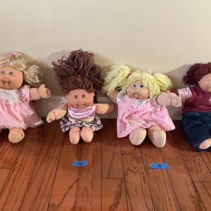 Peut inclure: Quatre poupées Cabbage Patch Kids vintage. Les poupées ont différentes couleurs de cheveux et tenues, dont des robes roses et des jeans en denim. Les poupées sont disposées sur un sol en bois.