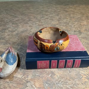 May include: A hand-painted ceramic ashtray with a brown and yellow design, resting on a book with a red spine. A small, carved duck figurine with blue and white paint is to the left of the ashtray.