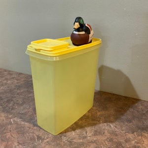 May include: A tall, pale yellow plastic container with a matching lid. A decorative duck figurine with brown, black, and white details sits on top. The container is rectangular and appears to be for storage.