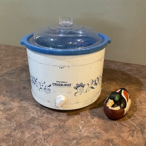 May include: A white Rival Crock-Pot stoneware slow cooker with a blue rim and a clear glass lid. The exterior features a blue floral pattern and the text Rival Crock-Pot Stoneware Slow Cooker with Off, Low, and High settings. A small duck figurine.