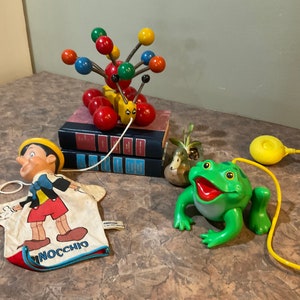 Könnte beinhalten: Eine Sammlung von Vintage-Spielzeug, darunter ein hölzernes Zugspielzeug mit bunten Kugeln, eine Pinocchio-Puppe und ein grünes Frosch-Zugspielzeug mit einer gelben Schnur.