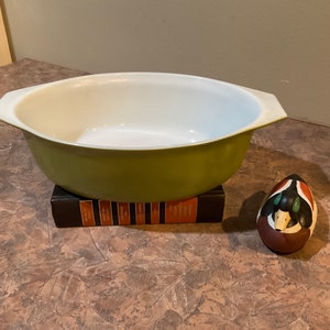 May include: A green oval baking dish with white interior. The dish is sitting on a stack of books. A wooden duck decoy is in the foreground.