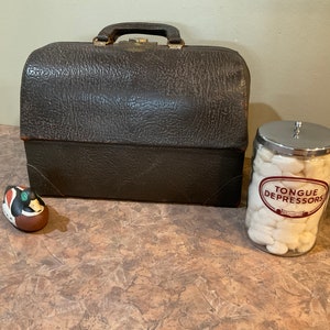 Może przedstawiać: Winietkowy brązowy skórzany kuferek lekarski z metalowym zapięciem. Szklany słoik ze srebrną pokrywką wypełniony białymi wacikami z etykietą "Tongue Depressors" stoi na brązowym blacie obok małej brązowo-zielonej figurki kaczki.