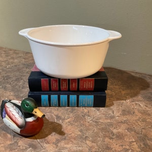 May include: White ceramic baking dish with two handles, sitting on top of a stack of three books. A painted ceramic duck figurine is in the foreground.