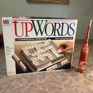May include: A vintage Milton Bradley Upwords game box with red and green lettering. The game board features letter tiles, and a wooden cat figurine is to the right. The box reads "A 3-DIMENSIONAL WORDGAME".