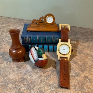 Könnte beinhalten: Eine Holzuhr mit einem braunen Lederarmband, eine hölzerne Entenfigur, eine Holzvase und zwei Bücher mit blauen Einbänden. Die Uhr hat ein weißes Zifferblatt mit schwarzen Zahlen und Zeigern.