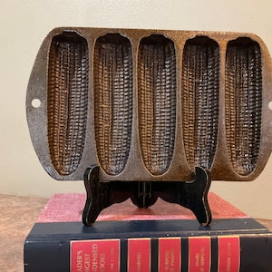 May include: A vintage cast iron cornbread pan with six individual moulds shaped like ears of corn. The pan is dark grey and has a rustic look.