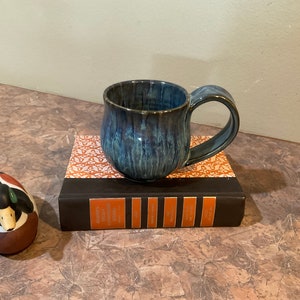 May include: A blue and green ceramic mug with a rounded body and a curved handle. The mug is sitting on top of a brown book with a patterned cover.
