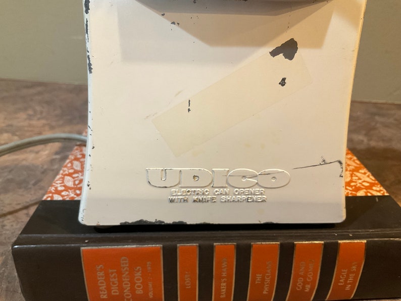 Peut inclure: Ouvre-bo&icirc;te &eacute;lectrique blanc avec un logo "UDICO" argent&eacute; et le texte "ELECTRIC CAN OPENER WITH KNIFE SHARPENER".
